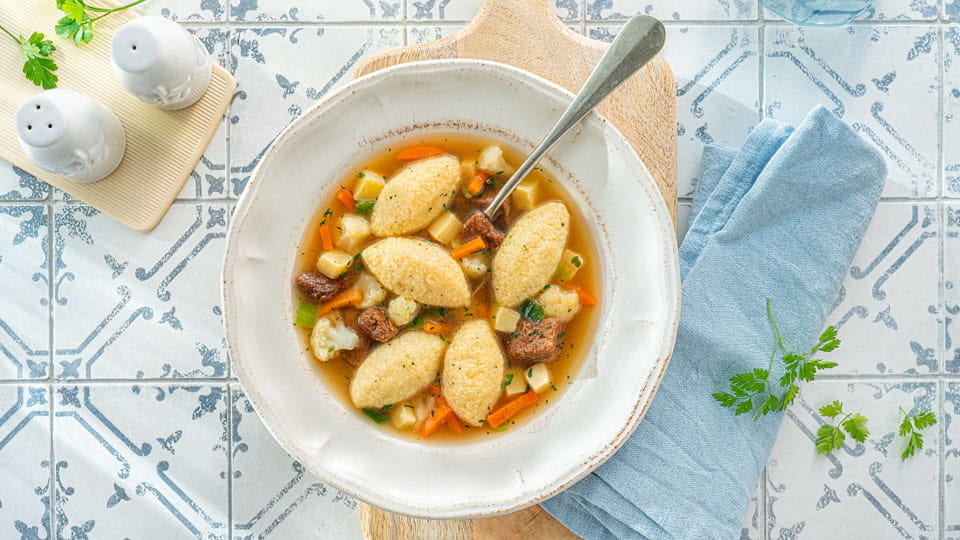 Tiefe Schale mit klarer Suppe, Grießnocken, Gemüsewürfeln und kleinen Fleischstücken, daneben hellblaue Serviette, Salz- und Pfefferstreuer auf gemustertem Fliesentisch.