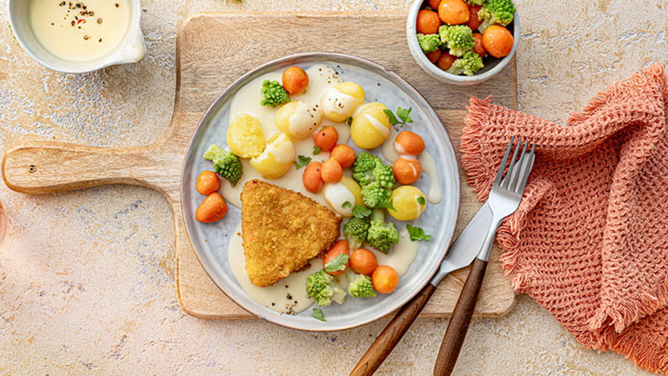 Runder Teller mit panierter Karottenecke, heller Soße, kleinen Kartoffeln, Karotten und grünen Romanesco-Röschen, auf Holzbrett mit orangefarbener Serviette und Besteck.