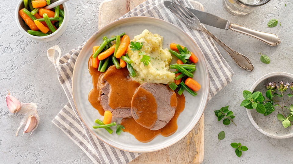 Teller mit zwei Scheiben Lammbraten in brauner Soße, Kartoffelpüree und Gemüse aus Karotten und grünen Bohnen, dekoriert mit Petersilie, auf grauem Untergrund mit Besteck und Kräutern.