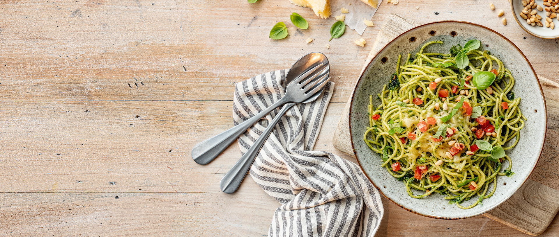 Spaghetti mit Pesto und Tomaten auf einem tiefen Teller. Daneben liegt Besteck auf einer Serviette auf einem hellen Holztisch
