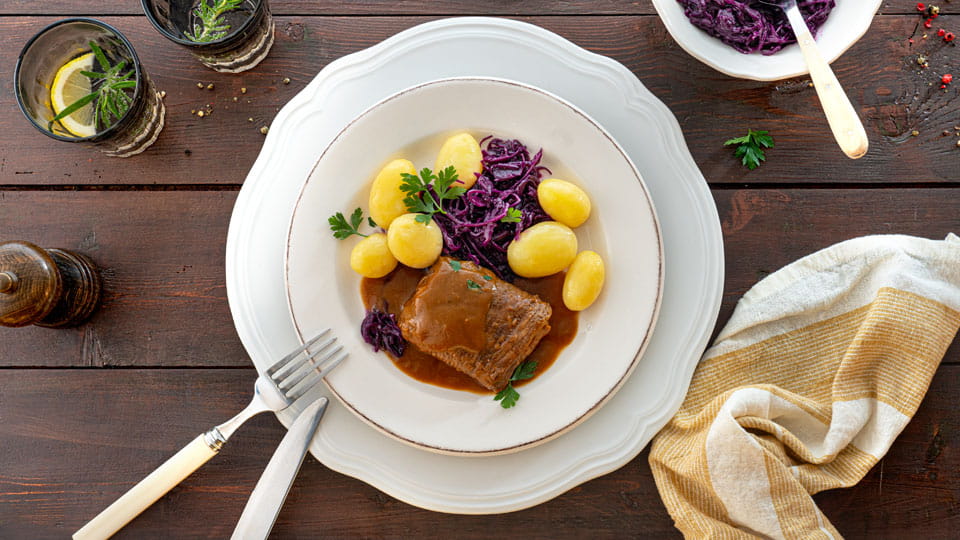 Teller mit Rinderroulade in dunkler Soße, kleinen Kartoffeln und Rotkohl, angerichtet auf dunklem Holztisch mit gelb-weißer Serviette und Glas mit Zitronenscheibe.