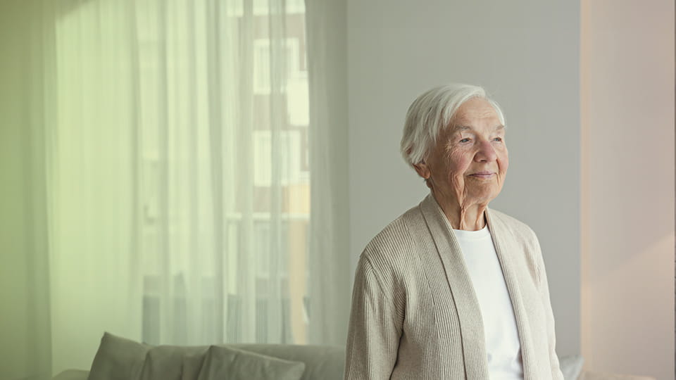 Seniorin steht lächelnd in einem hellen Wohnzimmer