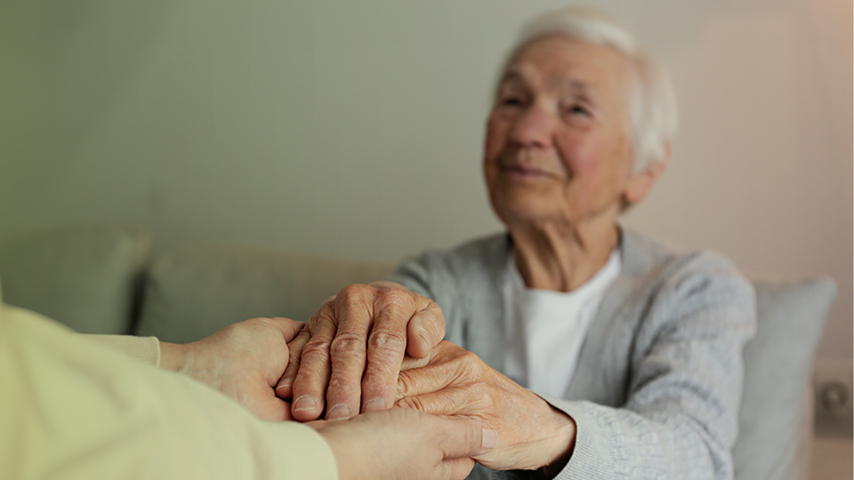 Seniorin hält die Hände einer anderen Person und lächelt dabei
