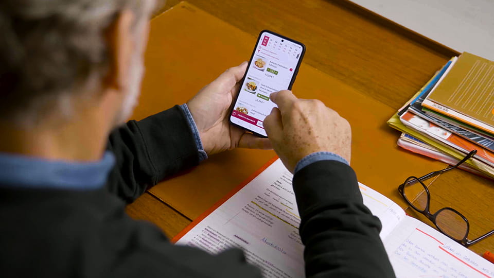 Mann sitzt an einem Schreibtisch und wählt auf seinem Smartphone ein Gericht aus der App aus