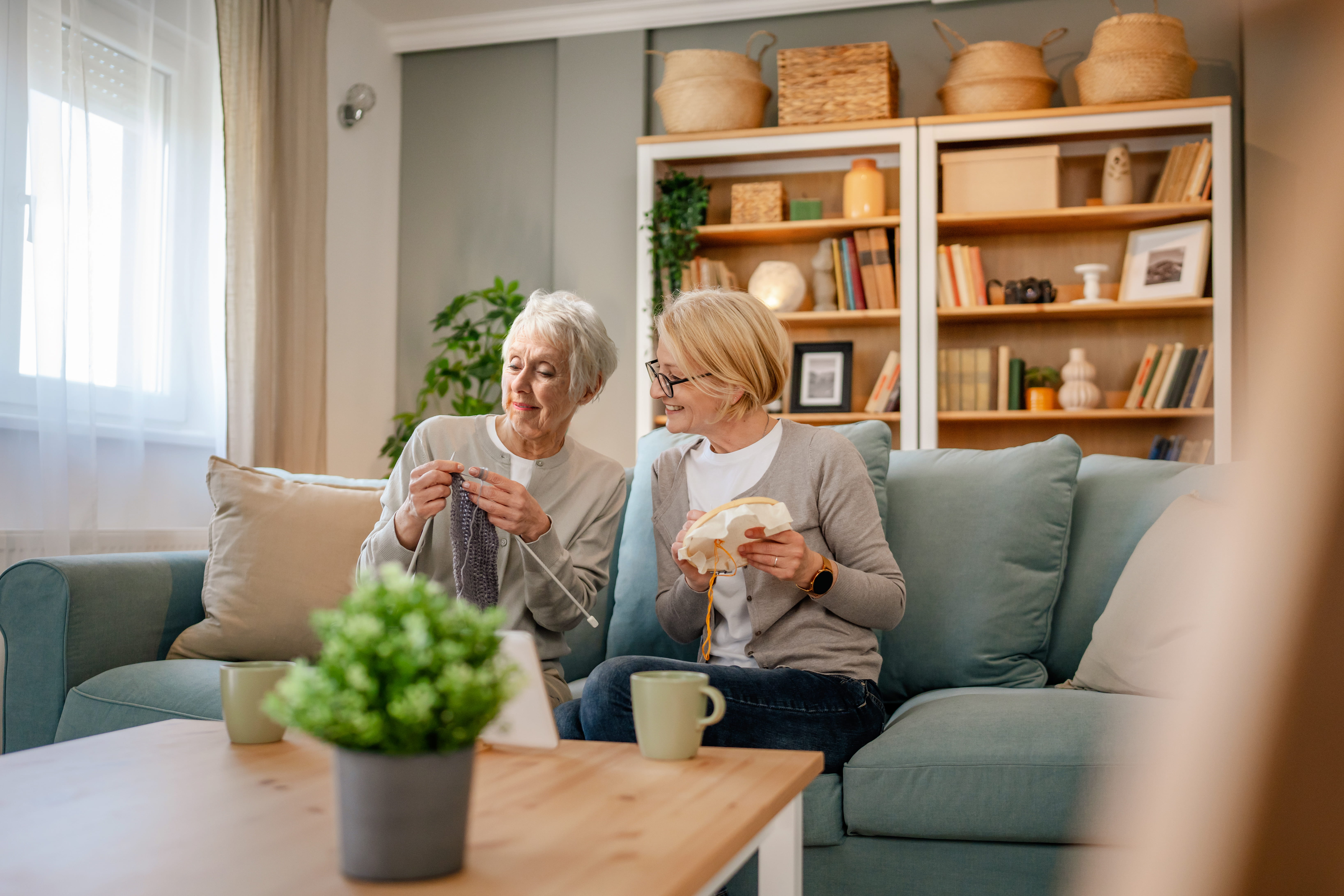 Frau und Seniorin sitzen im Wohnzimmer und stricken zusammen