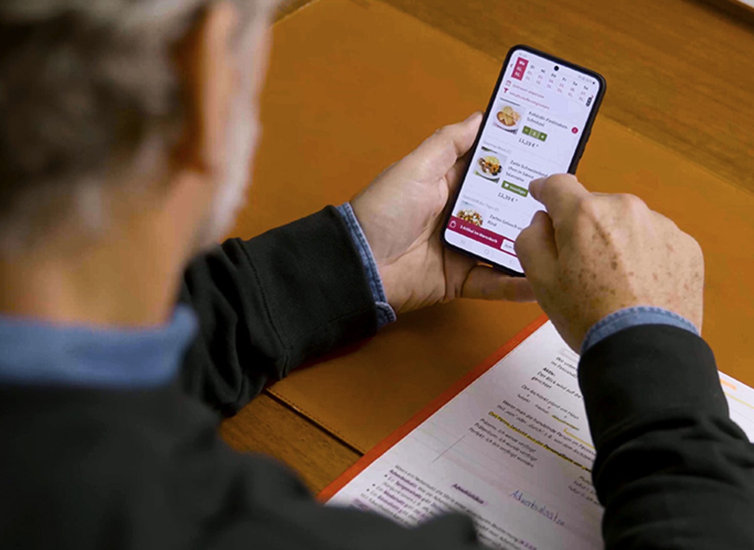 Mann sitzt an einem Schreibtisch und wählt auf seinem Smartphone ein Gericht aus der App aus