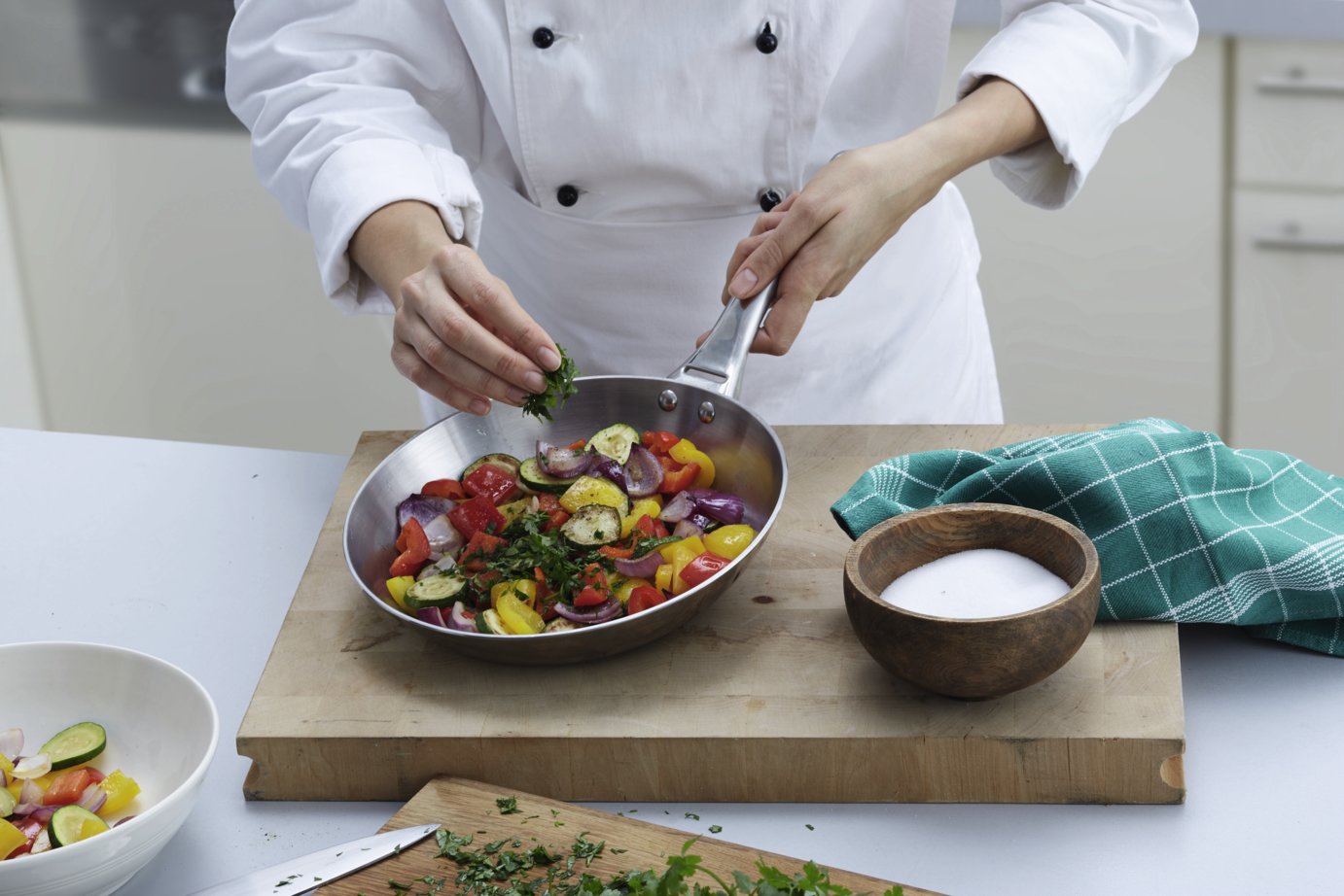 Koch in weißer Jacke bereitet in einer Pfanne Gemüse mit frischen Kräutern und Gewürzen zu