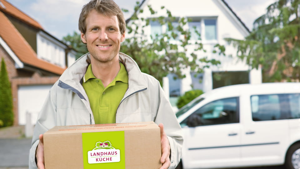 Lächelnder Kurier mit Landhausküche-Box vor einem weißen Lieferwagen