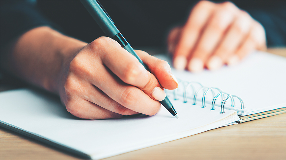 Eine Hand schreibt mit einem Stift in ein aufgeschlagenes Notizbuch