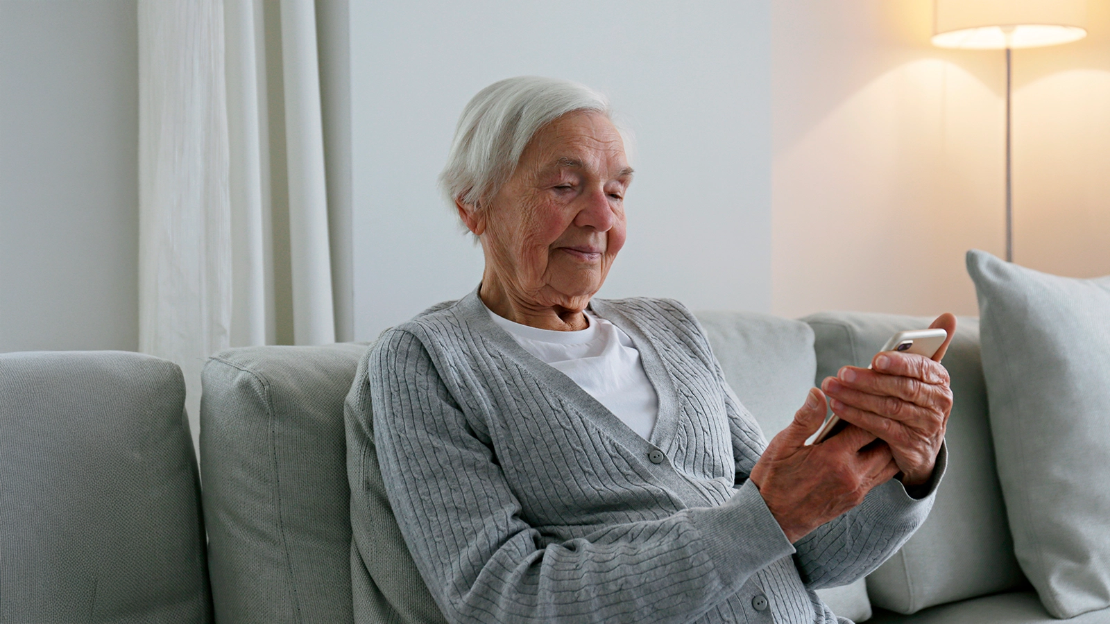 Eine Seniorin sitzt auf einem Sofa und hält ein Handy in der Hand und schaut drauf.