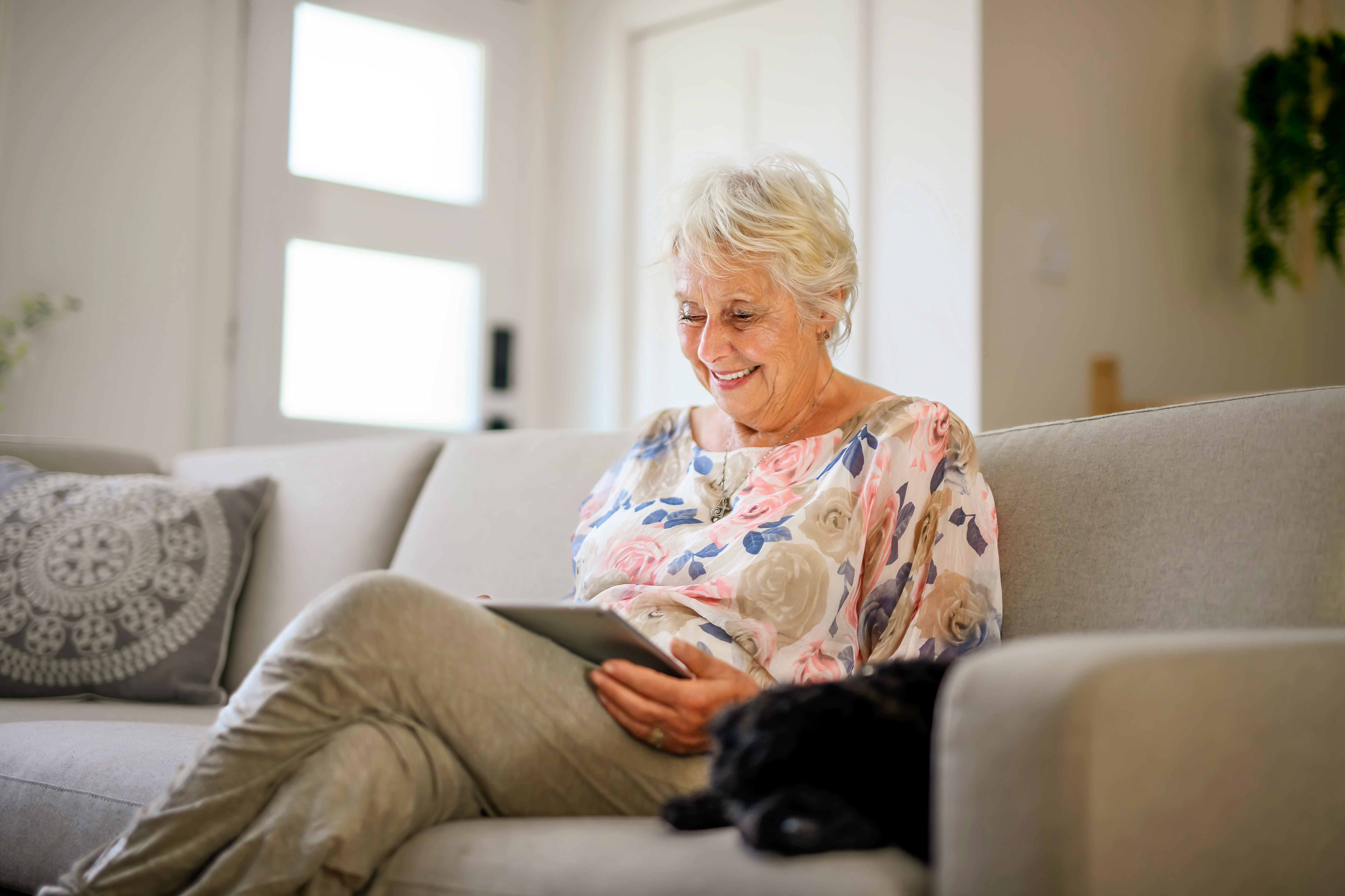 Seniorin sitzt auf dem Sofa, schaut auf ihr Tablet und lächelt dabei