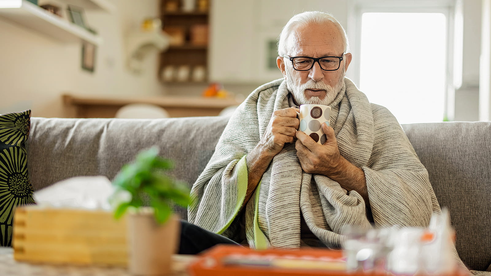 Ein älterer Mann sitzt krank auf dem Sofa und hält eine Tasse Tee in den Händen.