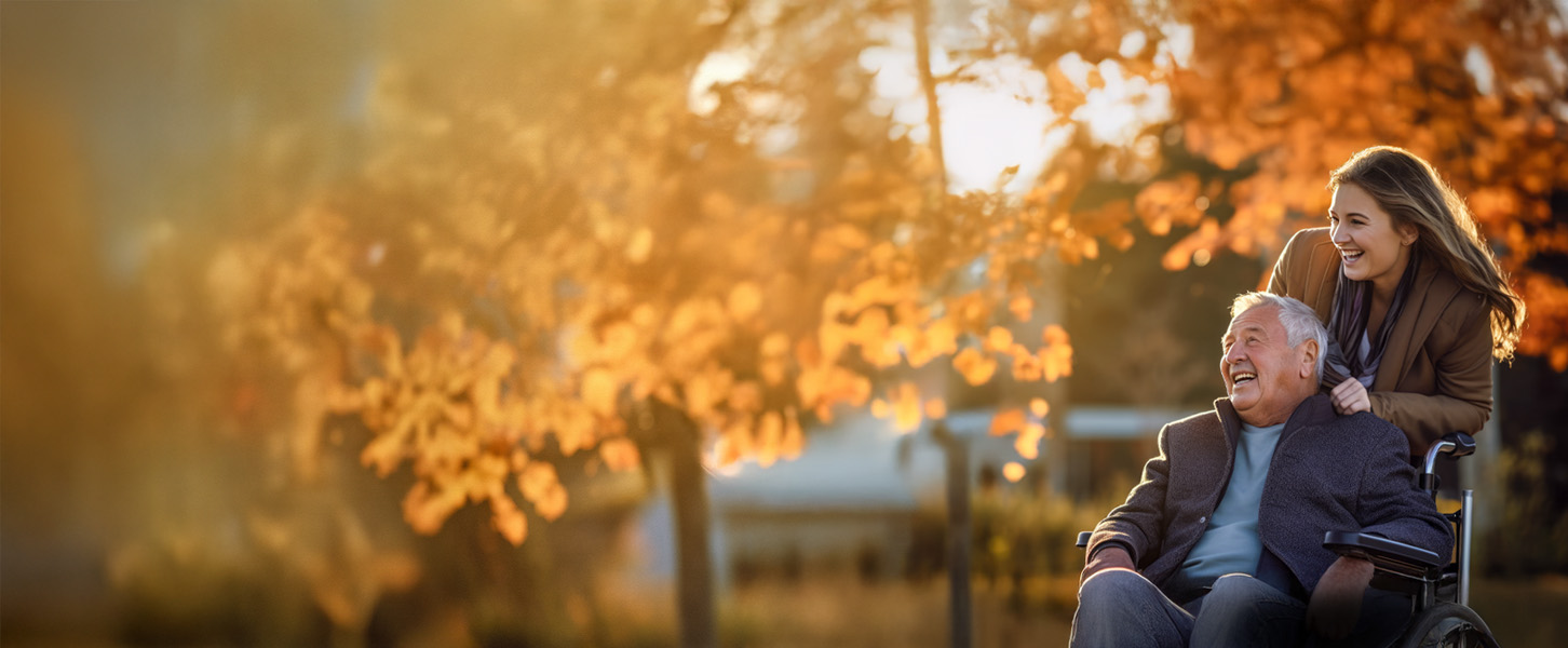 Ein im Rollstuhl sitzender Senior ist mit seiner Tochter an einem Herbsttag draußen im Park und lächelt.