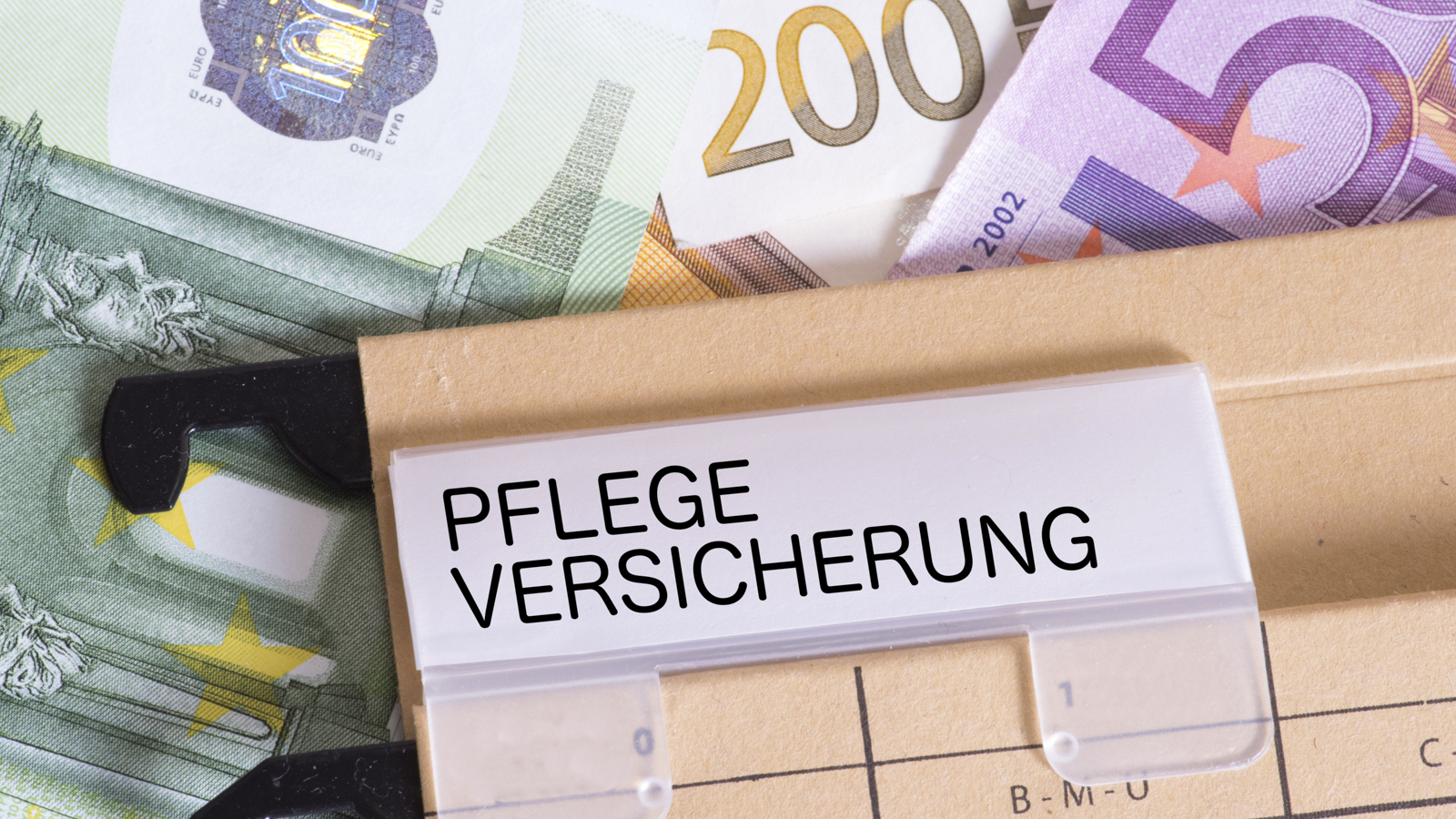 Ein Aktenorder mit der Aufschrift Pflegeversicherung und Gelscheine im Hintergrund.