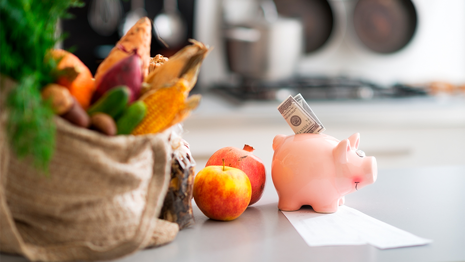 Einkaufstasche mit Obst und Gemüse, daneben ein Sparschwein mit Geldschein