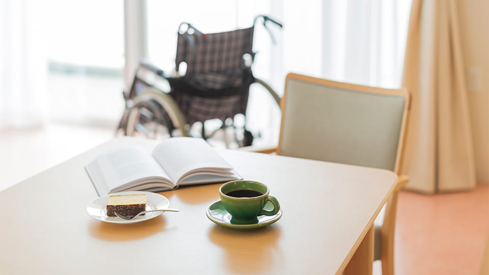 Tisch mit aufgeschlagenem Buch, Kaffee und Kuchen, im Hintergrund ein leerer Rollstuhl