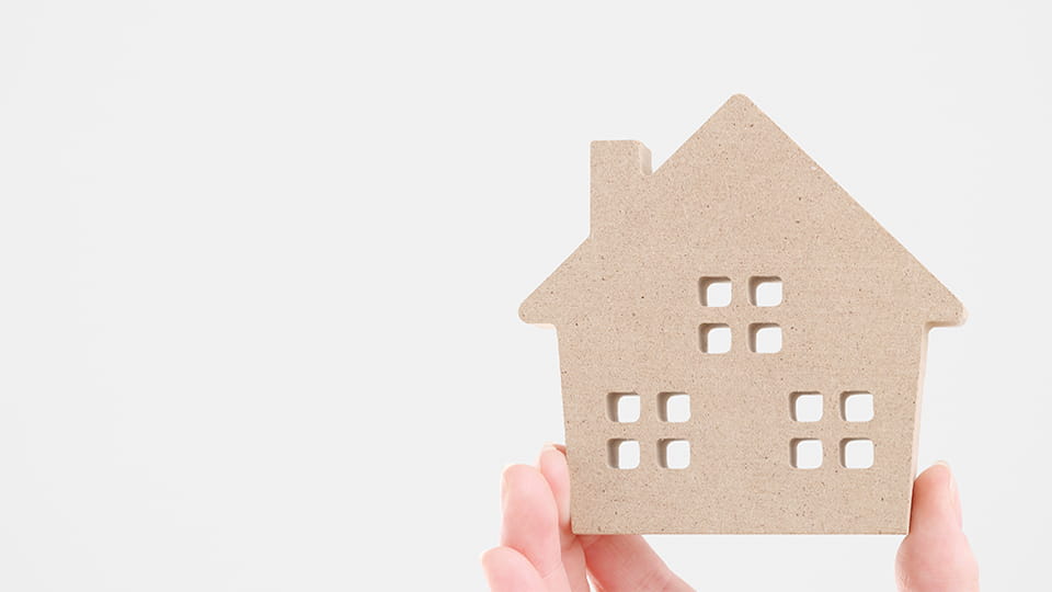 Abbildung eines Haus aus Pappe, welches von einer Hand hochgehalten wird