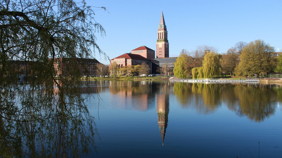 Blick über einen ruhigen See auf das Kieler Rathaus mit seinem hohen Turm, eingerahmt von Bäumen im Sonnenlicht