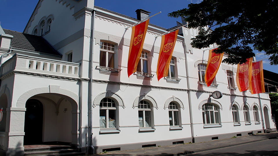 Das weiße Rathaus von Meerbusch mit roten Fahnen und von der Sonne angestrahlt