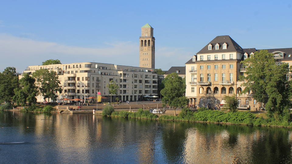 Blick auf Mülheim mit historischem Turm, modernen und alten Gebäuden am Ufer, davor ein Fluss