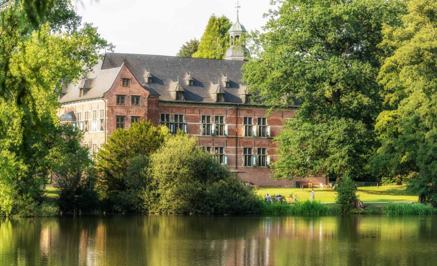 Das Schloss Reinbek mit Turm, umgeben von großen Bäumen und einem ruhigen See im Vordergrund