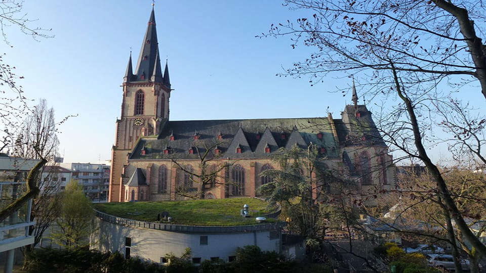 Apostelkirche mit hohem Turm und grüner Dachfläche im Vordergrund, umgeben von Bäumen und Wohnhäusern